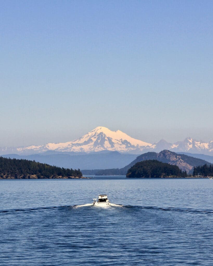 A white motorboat leaving a gentle wake as it cruises through calm blue waters, surrounded by forested islands, with snow-covered Mount Baker and the Cascade Range rising in the background under a clear sky.