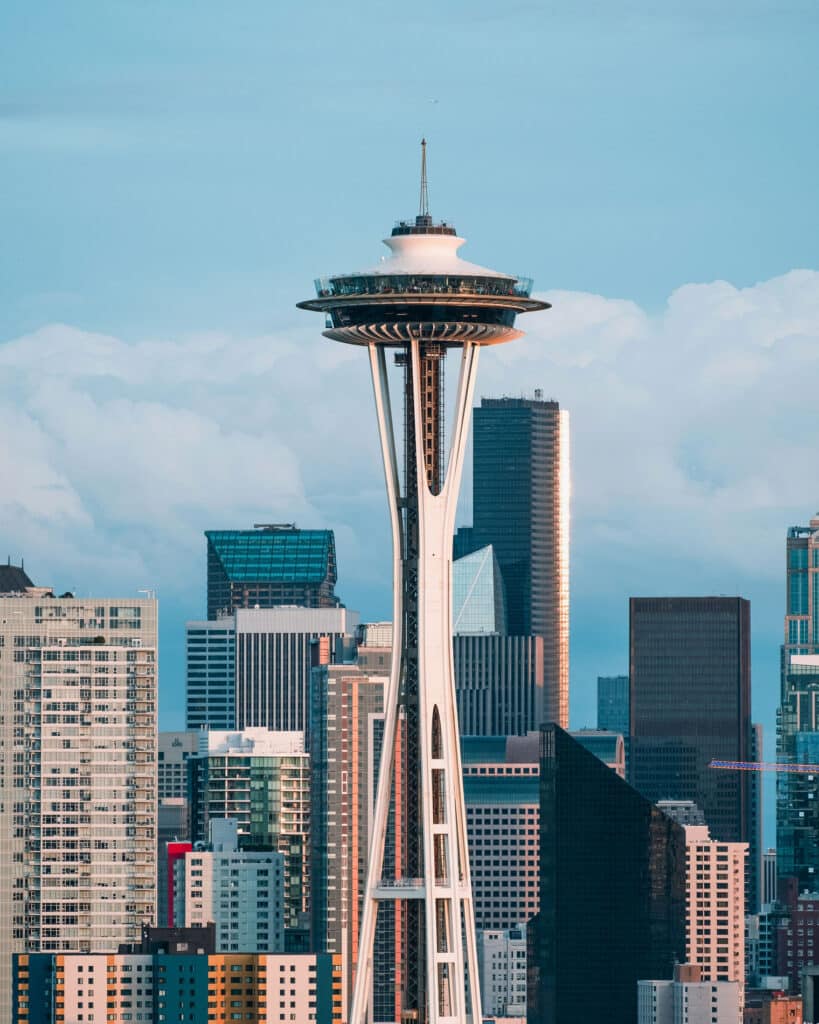 Detailed view of the Space Needle against a backdrop of downtown Seattle’s high-rise buildings, with soft blue skies and fluffy white clouds framing the iconic structure.